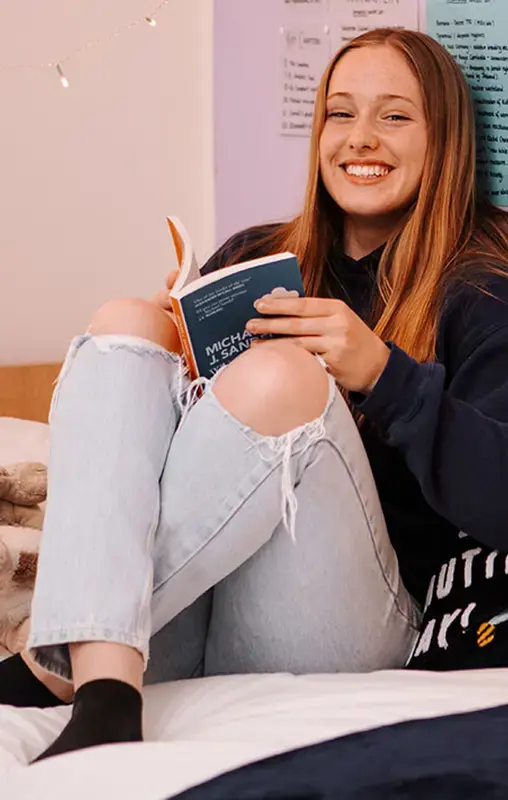 Gordonstoun pupil in the boarding house sitting on their bed, reading a book & smiling.