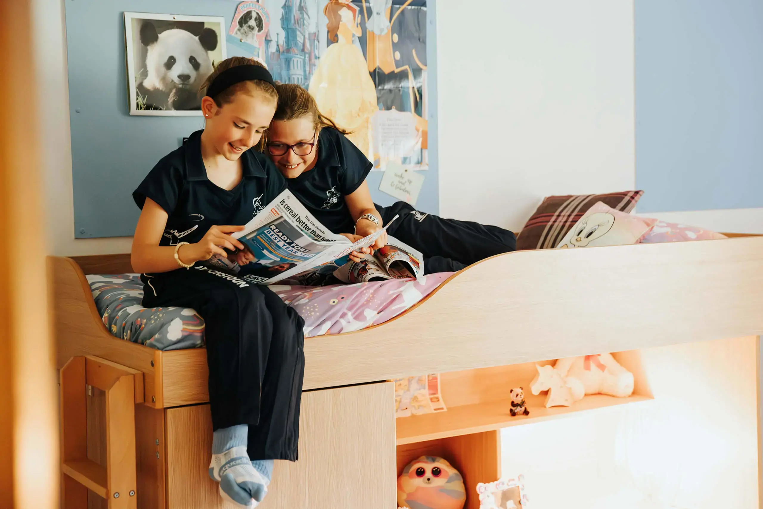 Prep aged girls sitting on a bed reading magazines and laughing.