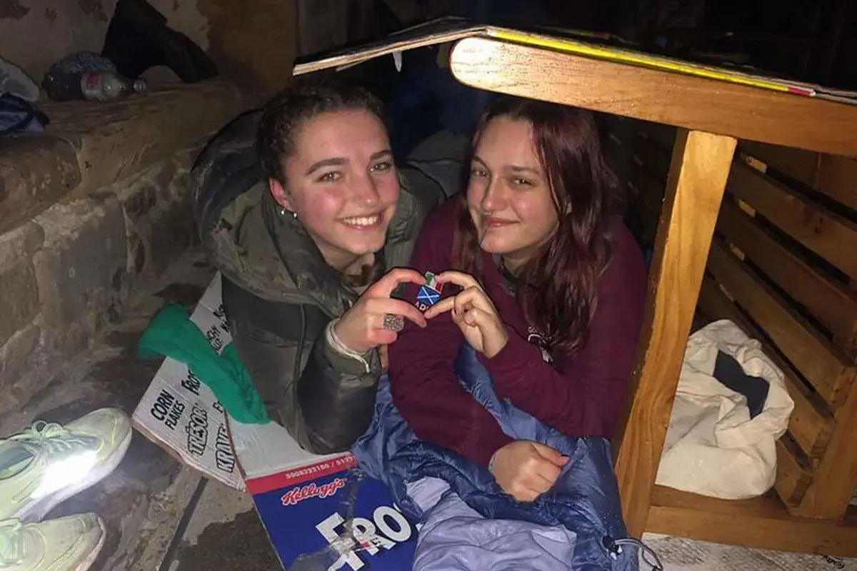 Huddled together, 2 students using a bench for shelter